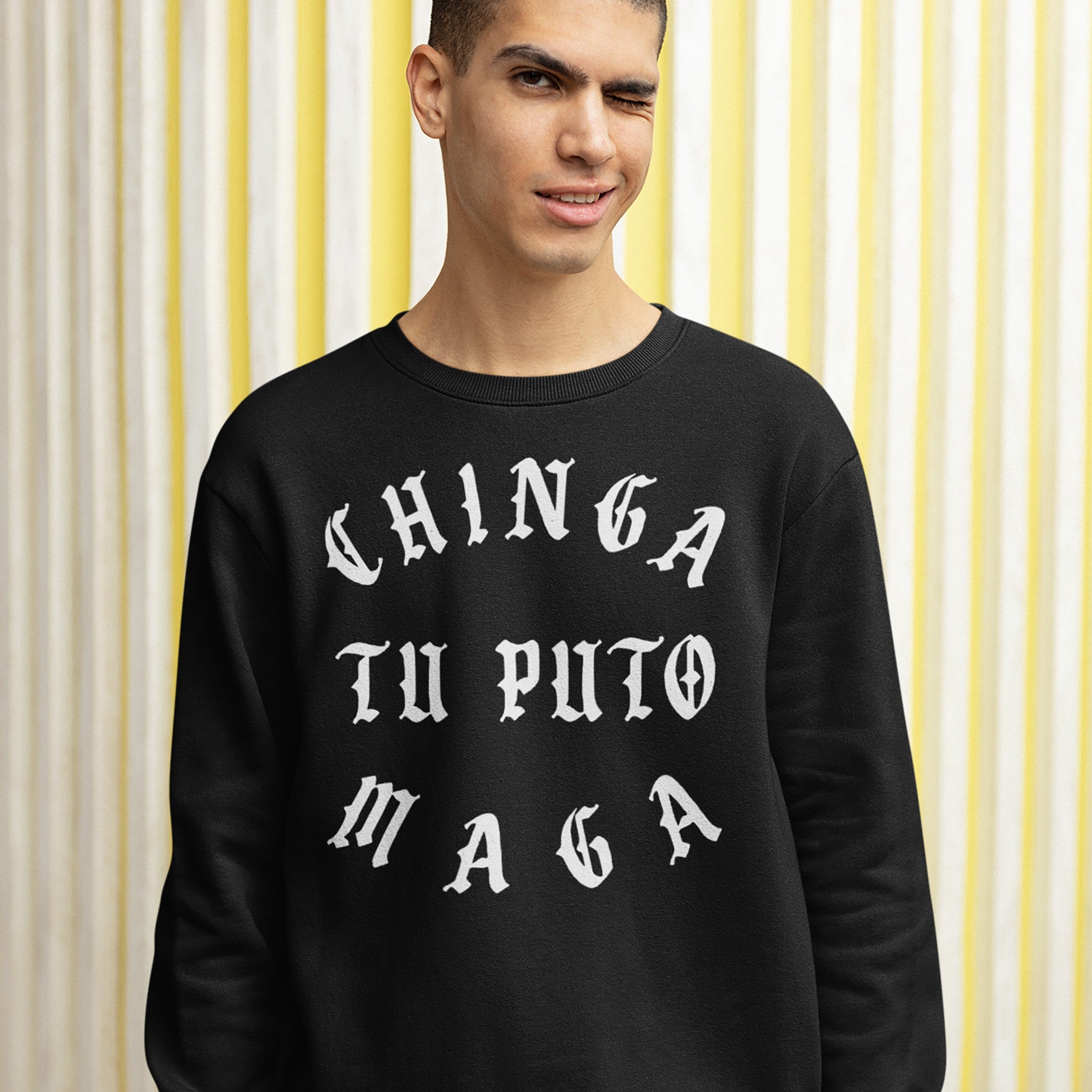 Chinga Tu MAGA Hoodie & Sweatshirt features bold, white Gothic-style text on a black sweatshirt. A young man winks at the camera, standing against a yellow-and-white backdrop, embodying a defiant, anti-fascist message.