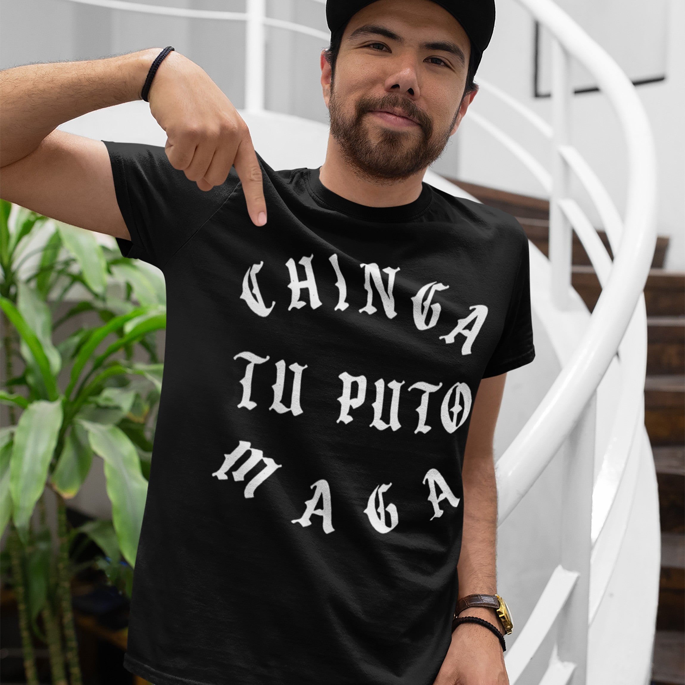 Man wearing the Chinga Tu MAGA T-shirt with bold white Gothic letters, expressing anti-fascist sentiment. He's pointing at the shirt, standing indoors near a spiral staircase and potted plants.