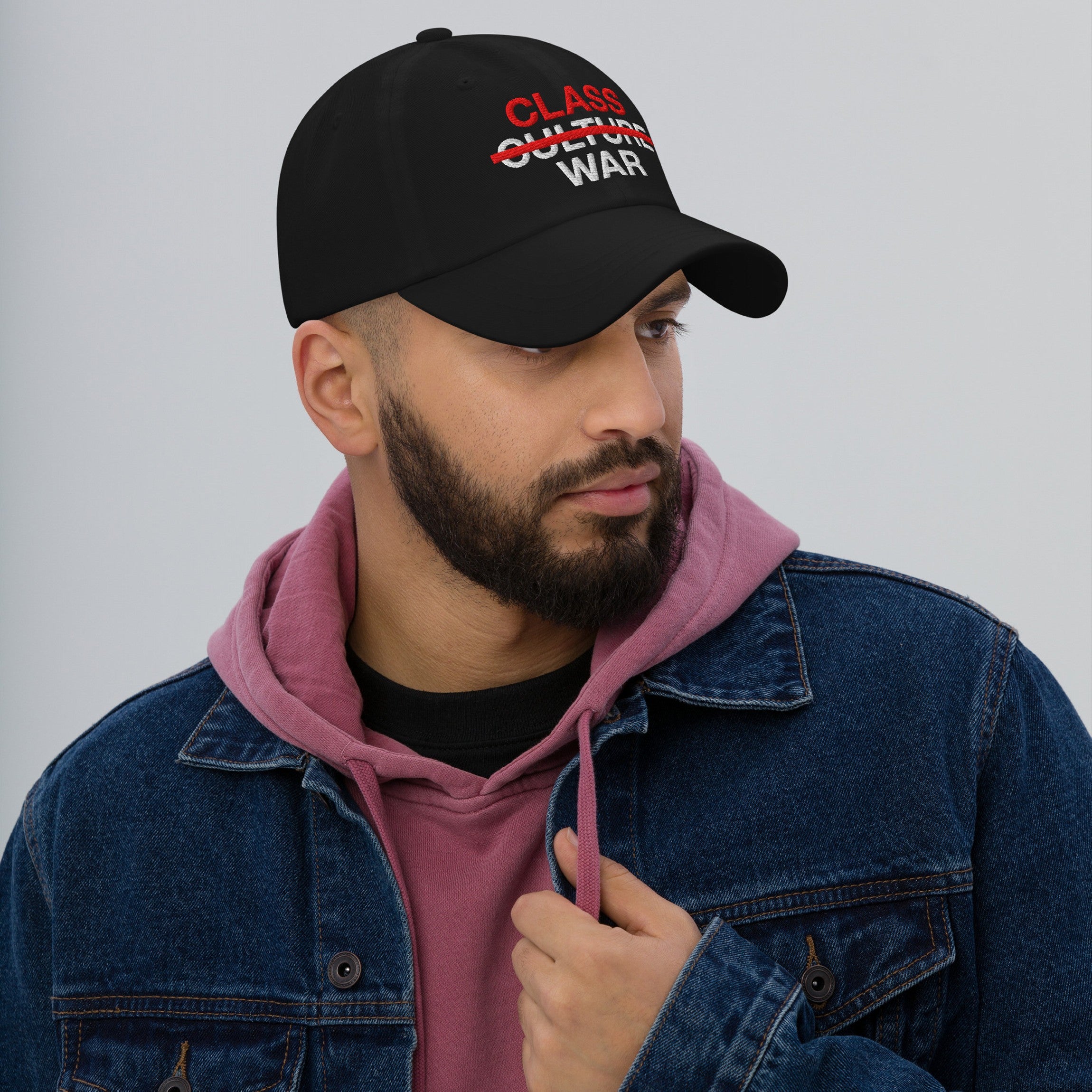 A bearded man models the Class War Hat, featuring bold red and white text highlighting economic justice by crossing out Culture War, emphasizing unity with a denim jacket and hoodie ensemble.