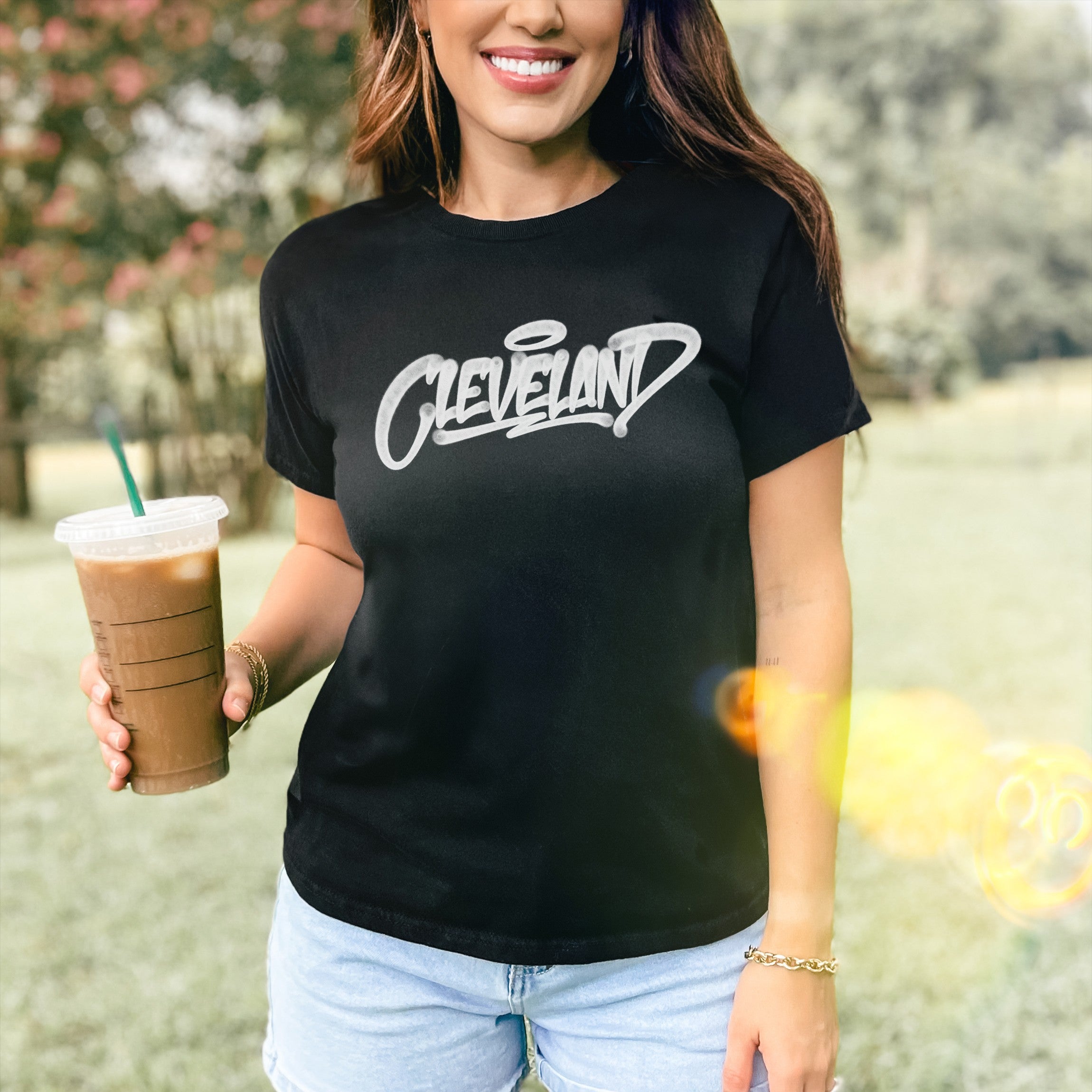 A woman in black Cleveland Handstyle Women's Baby Tee, showcasing white graffiti script, stands casually holding iced coffee, embodying a retro, fitted style with light-wash denim shorts and a gold bracelet.