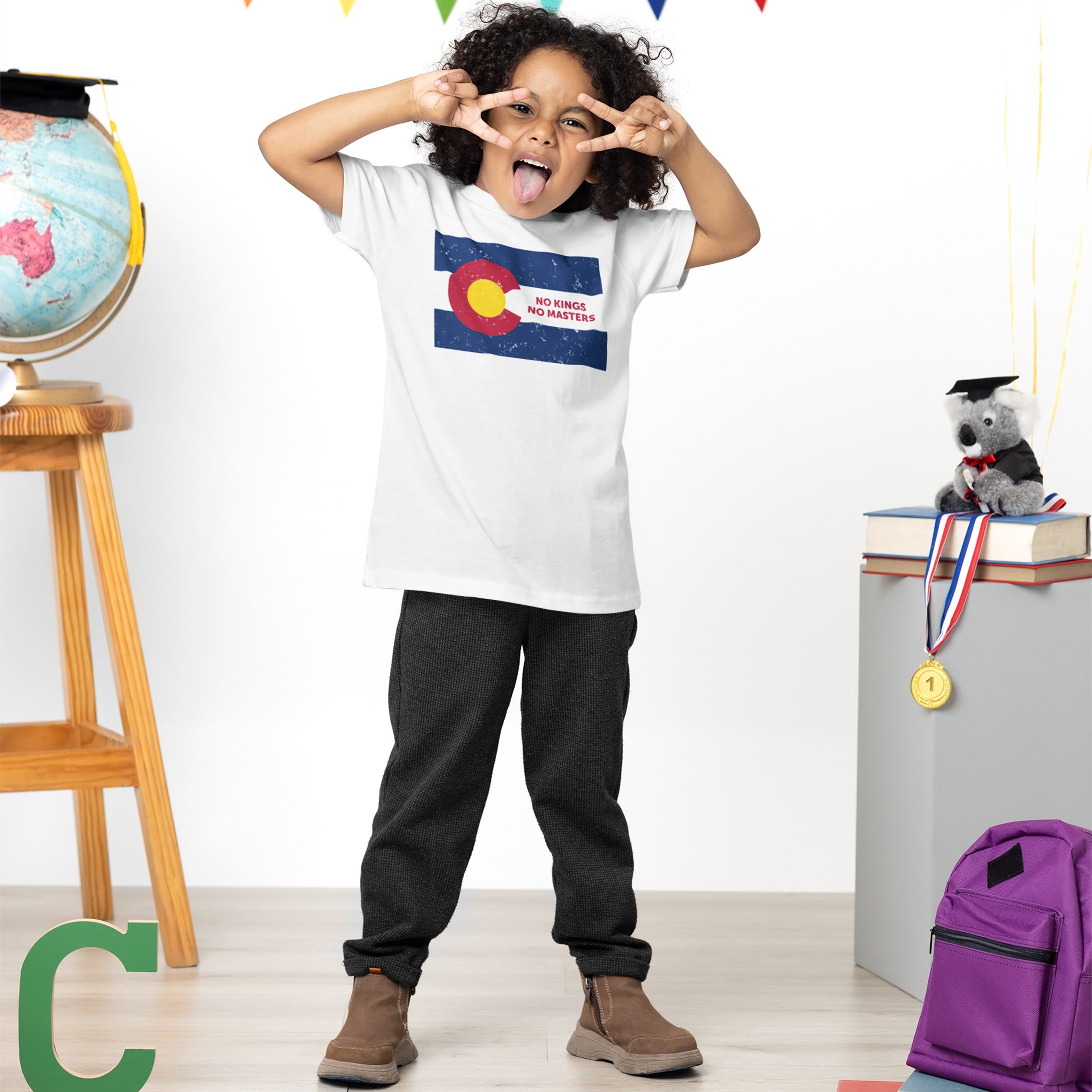 Young child in a Colorado No Kings Flag Shirt playfully poses indoors, surrounded by symbolic items like a globe and books, embodying a rebellious spirit and advocacy for autonomy.
