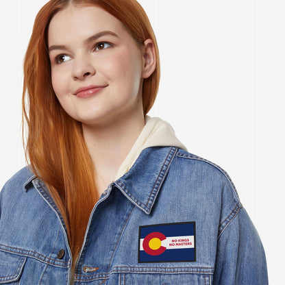 Young woman wears a light wash denim jacket featuring the Colorado Flag No Kings Patch, symbolizing self-governance with NO KINGS NO MASTERS slogan, against a plain white background.