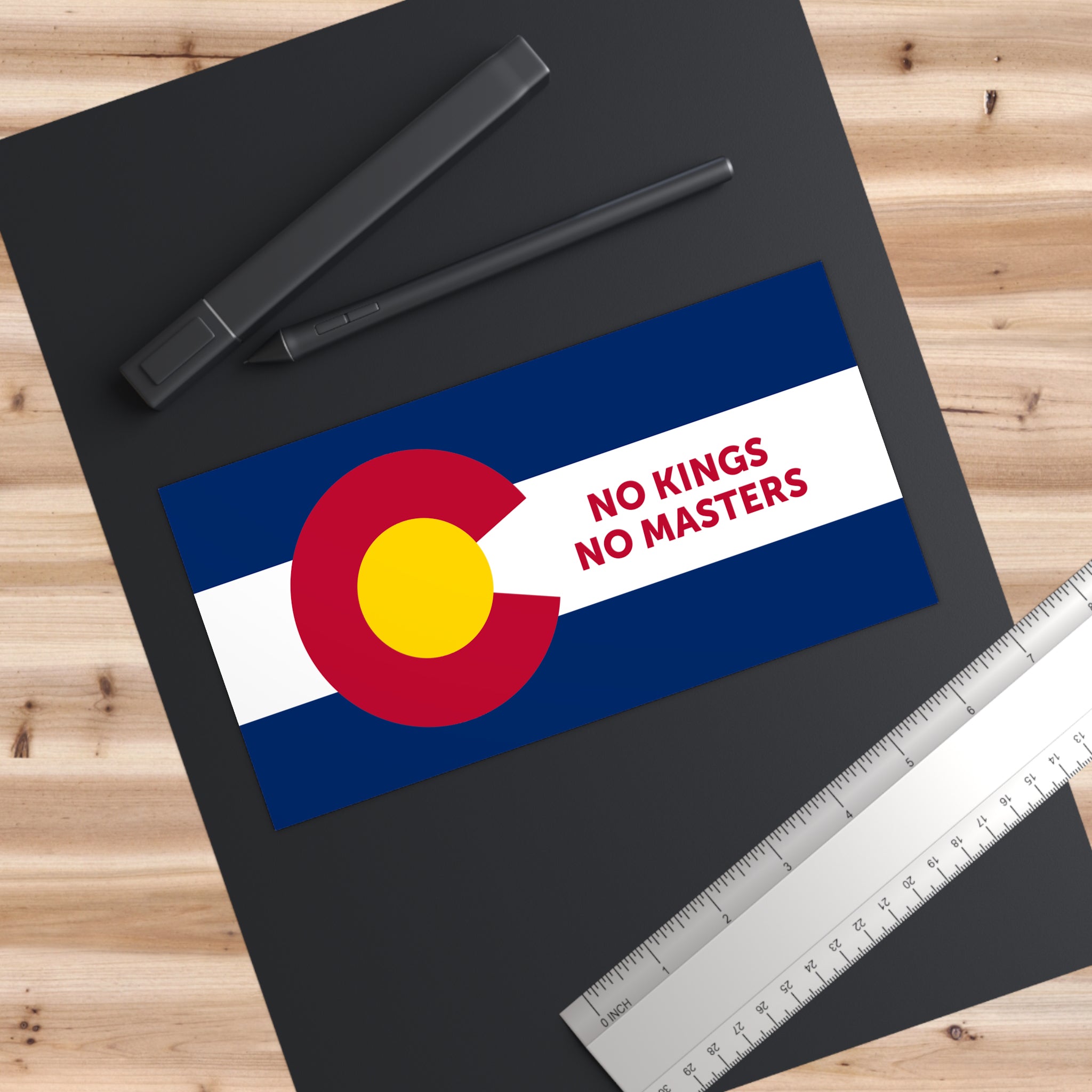 Colorado Flag No Kings Sticker & Magnet featuring a stylized state flag and slogan, positioned on a dark mat with a digital pen, capped marker, and metal ruler alongside.