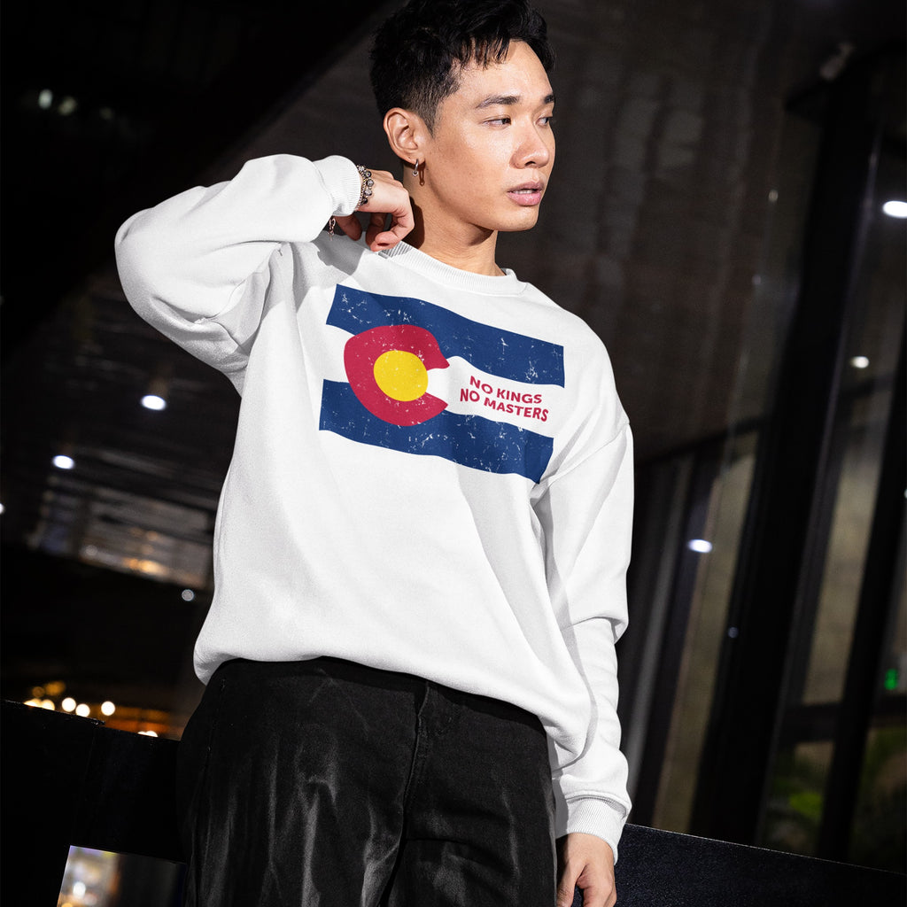 Young man in an atrium wearing a Colorado No Kings Flag Shirt, featuring a distressed Colorado flag graphic and the slogan “No Kings No Masters,” embodying rebellion and autonomy.