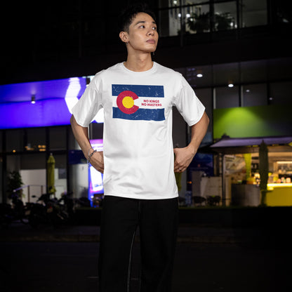 Young man in a Colorado No Kings Flag T-shirt, featuring a distressed state emblem and slogan, stands confidently outside a modern building, embodying a message of resistance and equality.