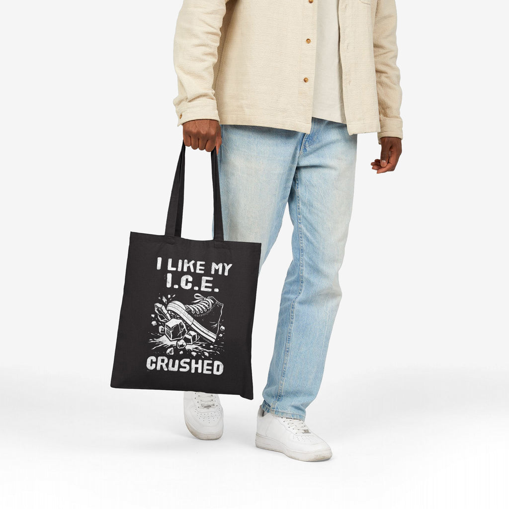 Person holds Crushed ICE Tote Bag, featuring a sneaker crushing ice cubes graphic and the slogan “I LIKE MY I.C.E. CRUSHED.”, symbolizing anti-fascist, pro-immigrant rights message on black canvas.
