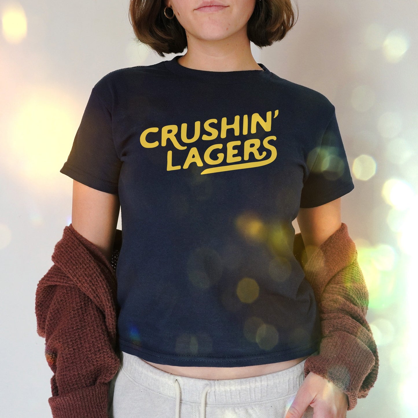 Person wearing the Crushin' Lagers Women's Baby Tee, featuring bold yellow lettering on navy fabric, paired with a maroon cardigan and gray sweatpants, embodying a relaxed, retro style.