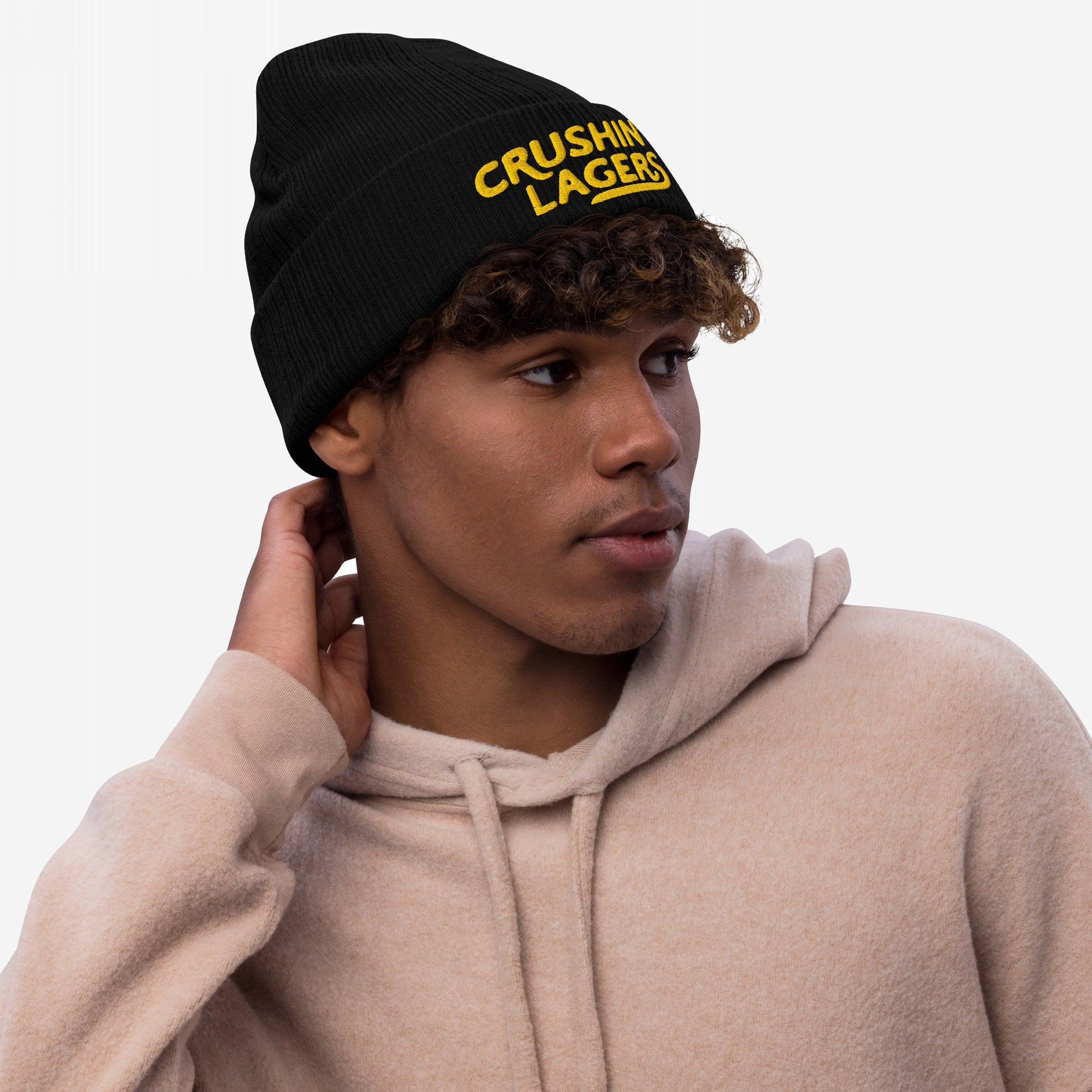 Young man wearing Crushin' Lagers Hat, a black knit beanie with yellow “CRUSHING LAGERS” text, paired with a light-tan hoodie. Perfect for lager enthusiasts, embodying casual style and good vibes.
