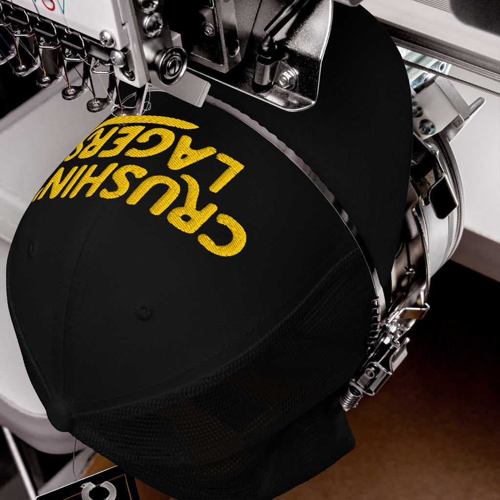Industrial embroidery machine stitching CRUSHIN' LAGERS onto a black baseball cap, showcasing the bold yellow letters on the curved crown. This cap celebrates the joy of lager and good vibes.