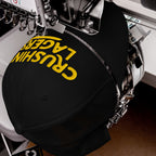 Industrial embroidery machine stitching CRUSHIN' LAGERS onto a black baseball cap, showcasing the bold yellow letters on the curved crown. This cap celebrates the joy of lager and good vibes.