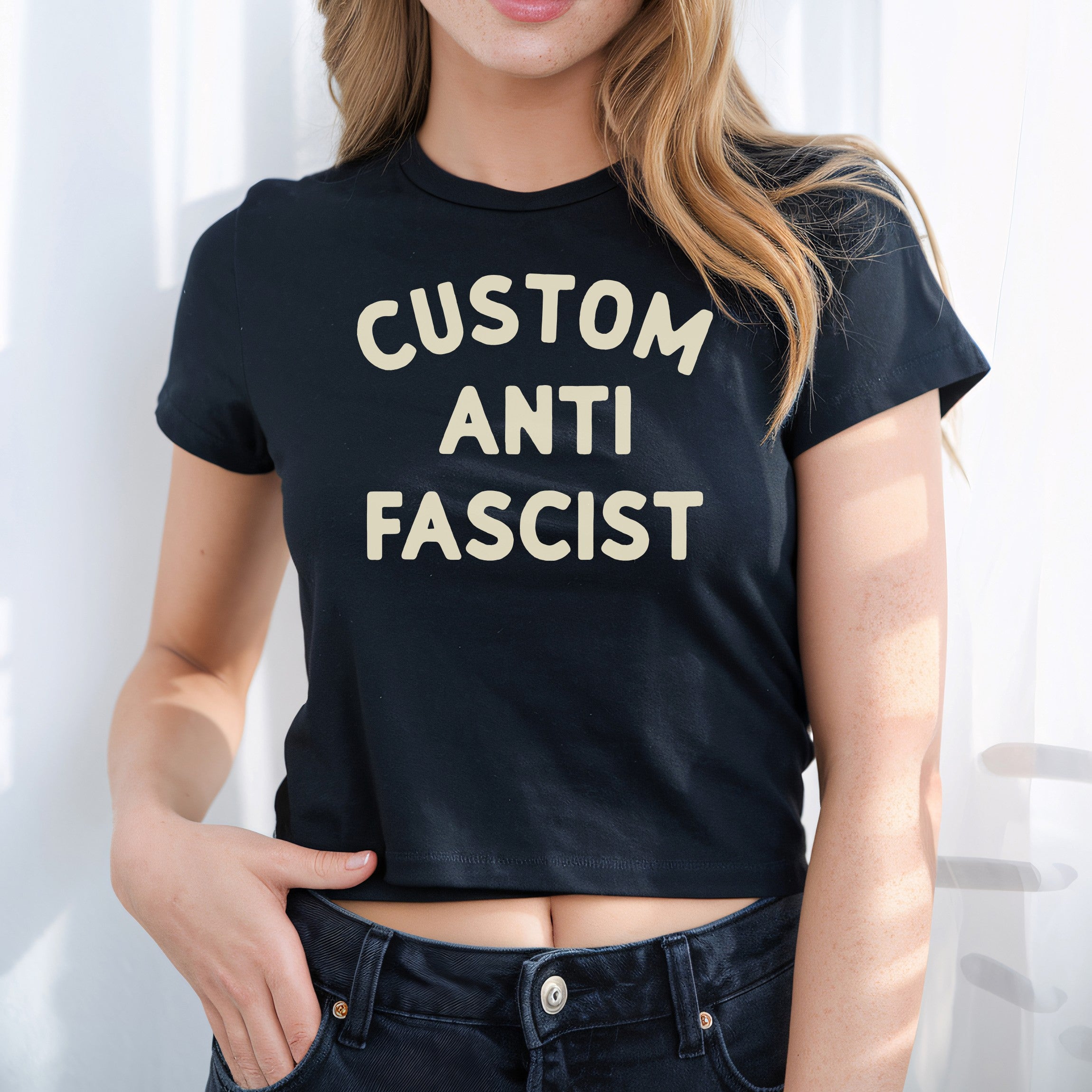 Young person wearing the Custom Anti-Fascist Women's Baby Tee, featuring bold cream text. This cropped black T-shirt reflects a retro Y2K style, paired with dark denim jeans, embodying activism and individuality.