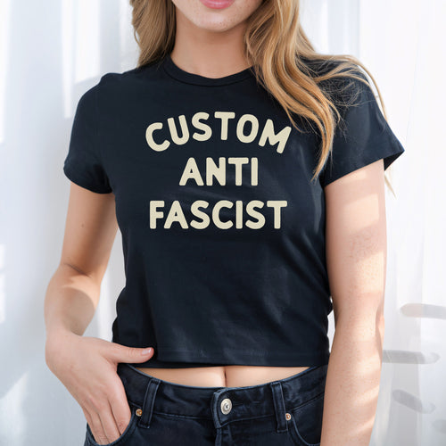 Young person wearing the Custom Anti-Fascist Women's Baby Tee, featuring bold cream text. This cropped black T-shirt reflects a retro Y2K style, paired with dark denim jeans, embodying activism and individuality.