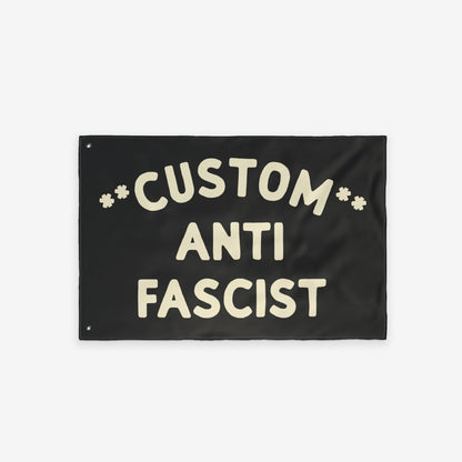 Custom Anti-Fascist Flag featuring bold, cream hand-drawn lettering with Custom arched above ANTI FASCIST and starburst symbols. Includes metal grommets for hanging. Suitable for personalizing with city or message.
