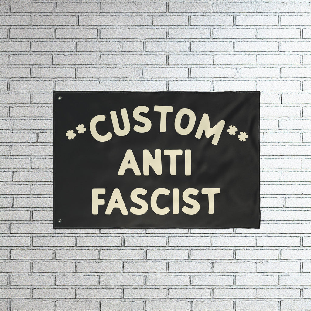 Custom Anti-Fascist Flag with bold off-white lettering on a black fabric, mounted on a white brick wall. This customizable flag powerfully communicates anti-fascist sentiment, ideal for rallies or home display.