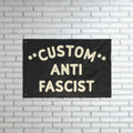 Custom Anti-Fascist Flag with bold off-white lettering on a black fabric, mounted on a white brick wall. This customizable flag powerfully communicates anti-fascist sentiment, ideal for rallies or home display.
