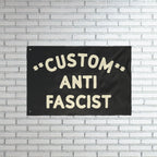 Custom Anti-Fascist Flag with bold off-white lettering on a black fabric, mounted on a white brick wall. This customizable flag powerfully communicates anti-fascist sentiment, ideal for rallies or home display.