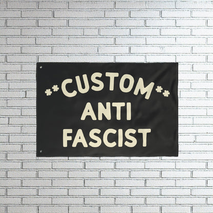 Custom Anti-Fascist Flag with bold off-white lettering on a black fabric, mounted on a white brick wall. This customizable flag powerfully communicates anti-fascist sentiment, ideal for rallies or home display.