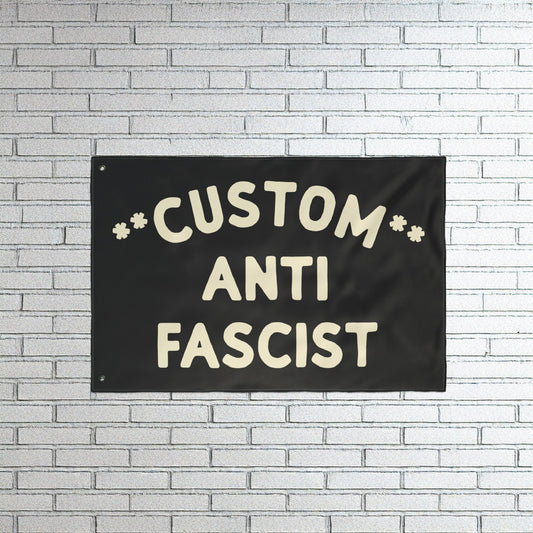 Custom Anti-Fascist Flag with bold off-white lettering on a black fabric, mounted on a white brick wall. This customizable flag powerfully communicates anti-fascist sentiment, ideal for rallies or home display.