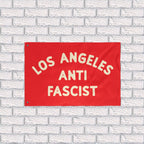 Custom Anti-Fascist Flag featuring LOS ANGELES ANTI FASCIST in bold cream text on a red background, mounted against a white brick wall. Durable polyester with metal grommets, ideal for rallies or home display.