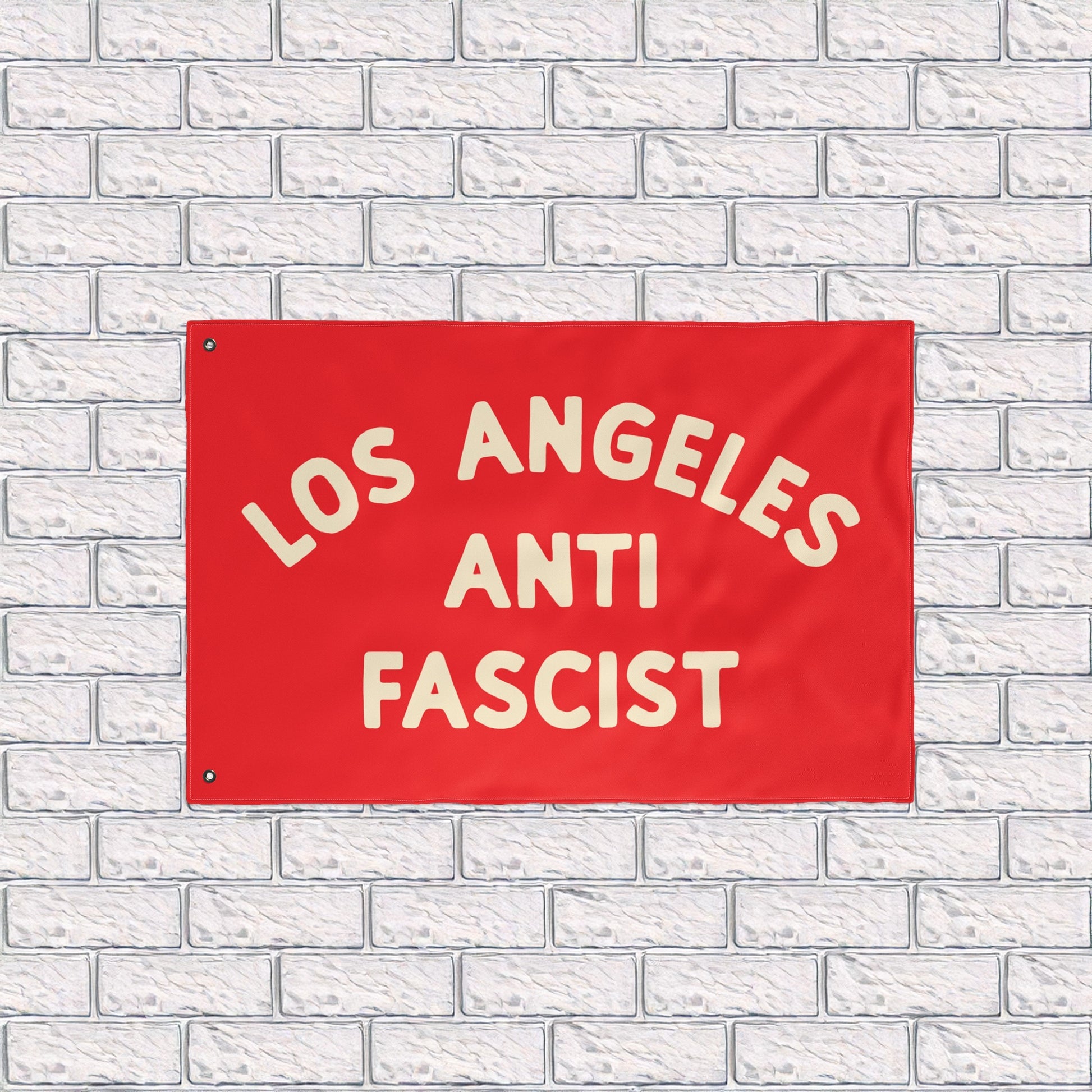 Custom Anti-Fascist Flag featuring LOS ANGELES ANTI FASCIST in bold cream text on a red background, mounted against a white brick wall. Durable polyester with metal grommets, ideal for rallies or home display.