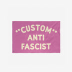 Custom Anti-Fascist Flag with bold cream letters on a pink background, featuring CUSTOM at the top, ANTI in the center, and FASCIST at the bottom, designed with two metal grommets for hanging.