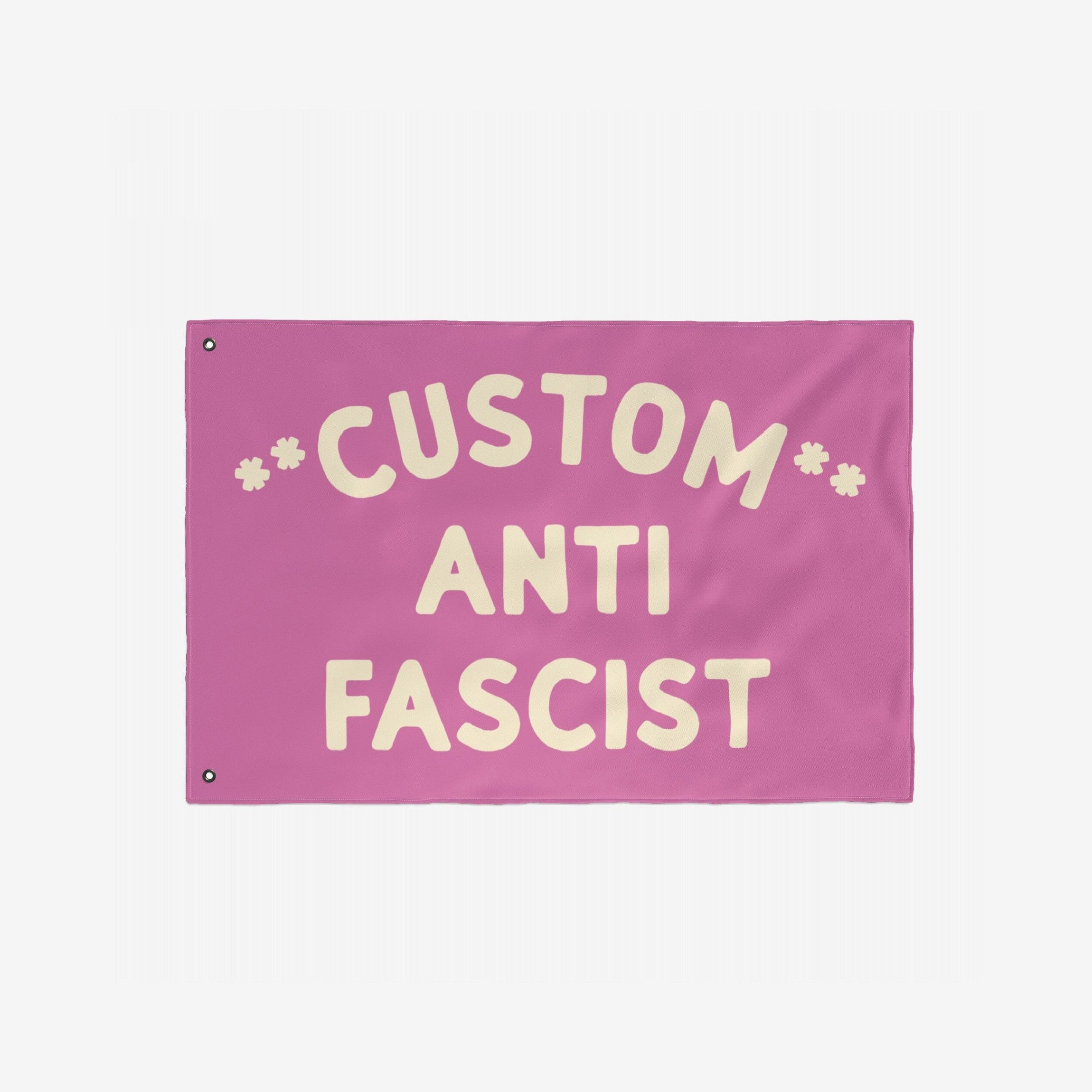 Custom Anti-Fascist Flag with bold cream letters on a pink background, featuring CUSTOM at the top, ANTI in the center, and FASCIST at the bottom, designed with two metal grommets for hanging.