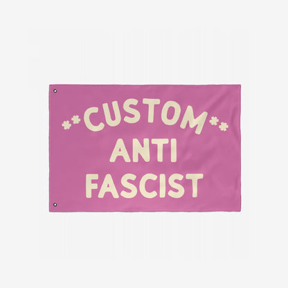 Custom Anti-Fascist Flag with bold cream letters on a pink background, featuring CUSTOM at the top, ANTI in the center, and FASCIST at the bottom, designed with two metal grommets for hanging.