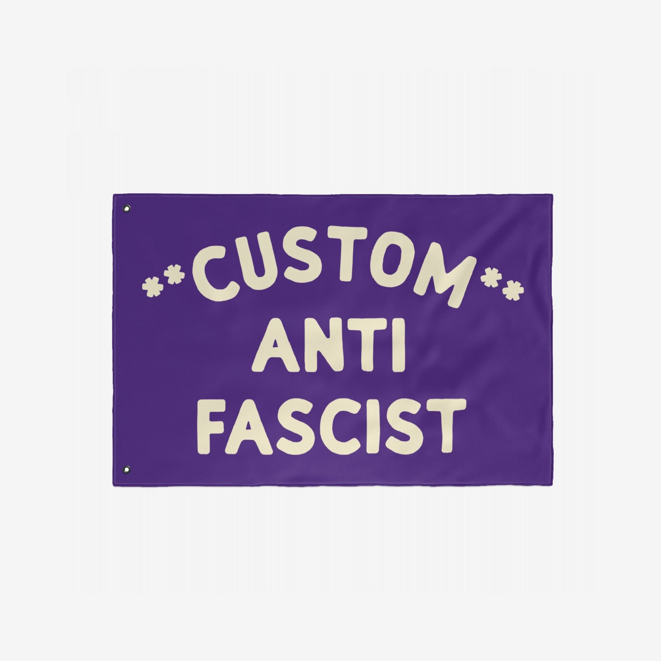 Custom Anti-Fascist Flag with hand-lettered text CUSTOM ANTI FASCIST and star-like asterisks. Features two grommets for hanging, symbolizing unity and resistance against fascism.