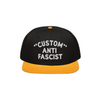 Custom Anti-Fascist Snapback Hat featuring a black crown and yellow brim, with bold white CUSTOM ANTI FASCIST embroidery. Per