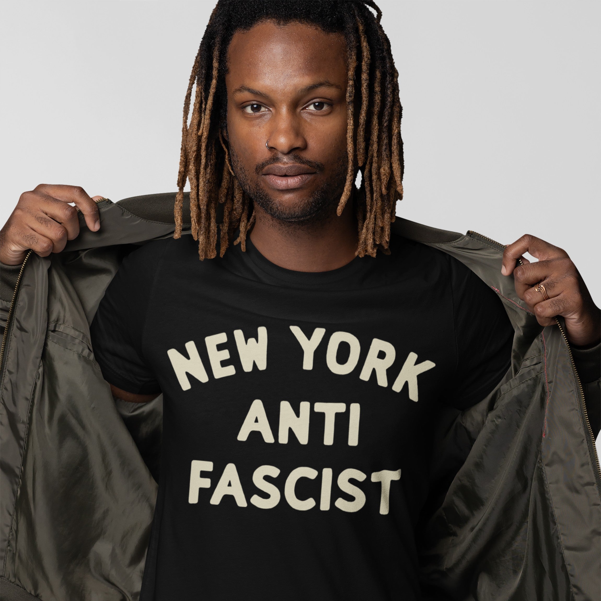 Man showcasing Custom Anti-Fascist T-shirt, featuring NEW YORK ANTI FASCIST slogan in off-white block letters, layered under a dark bomber jacket, combining activism with style.