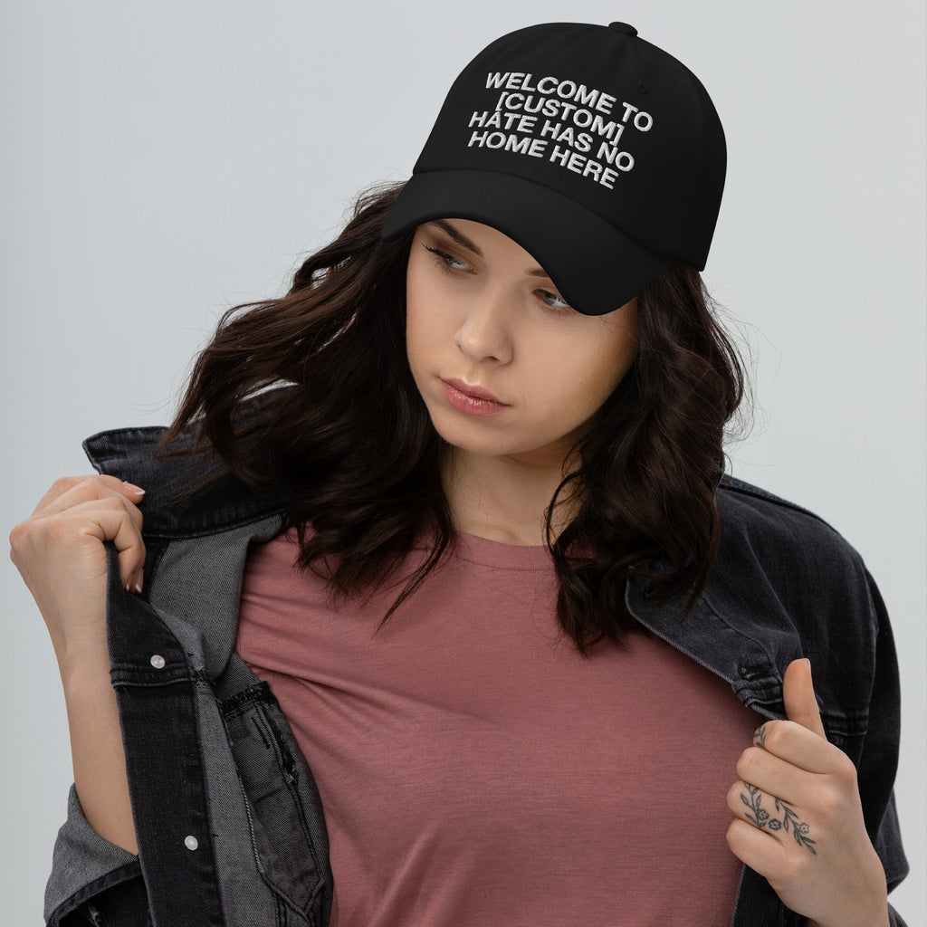 Young woman models the Custom Hate Has No Home Here Dad Hat, showcasing its embroidered message. She pairs it with a black denim jacket and mauve T-shirt, embodying inclusive style and activism.