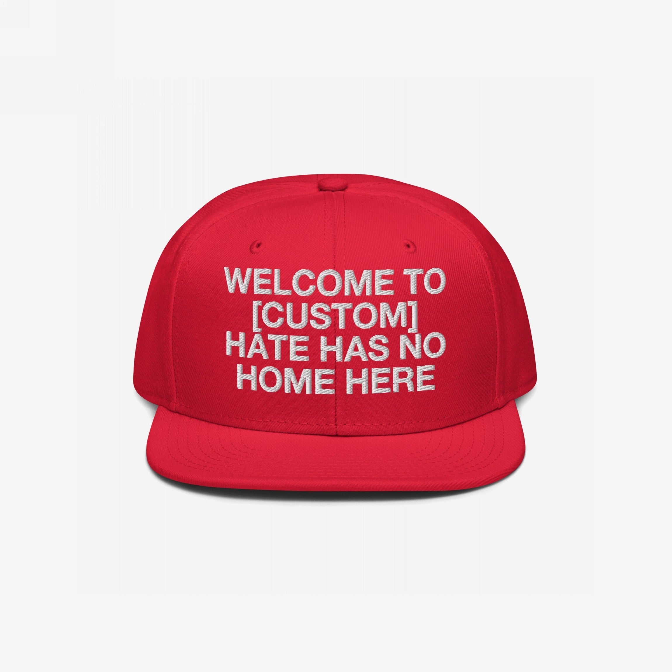 Red flat-brim cap with white embroidery reads WELCOME TO [CUSTOM] HATE HAS NO HOME HERE. Customizable snapback hat, promoting unity and anti-hate values, ideal for personalizing with your city name.