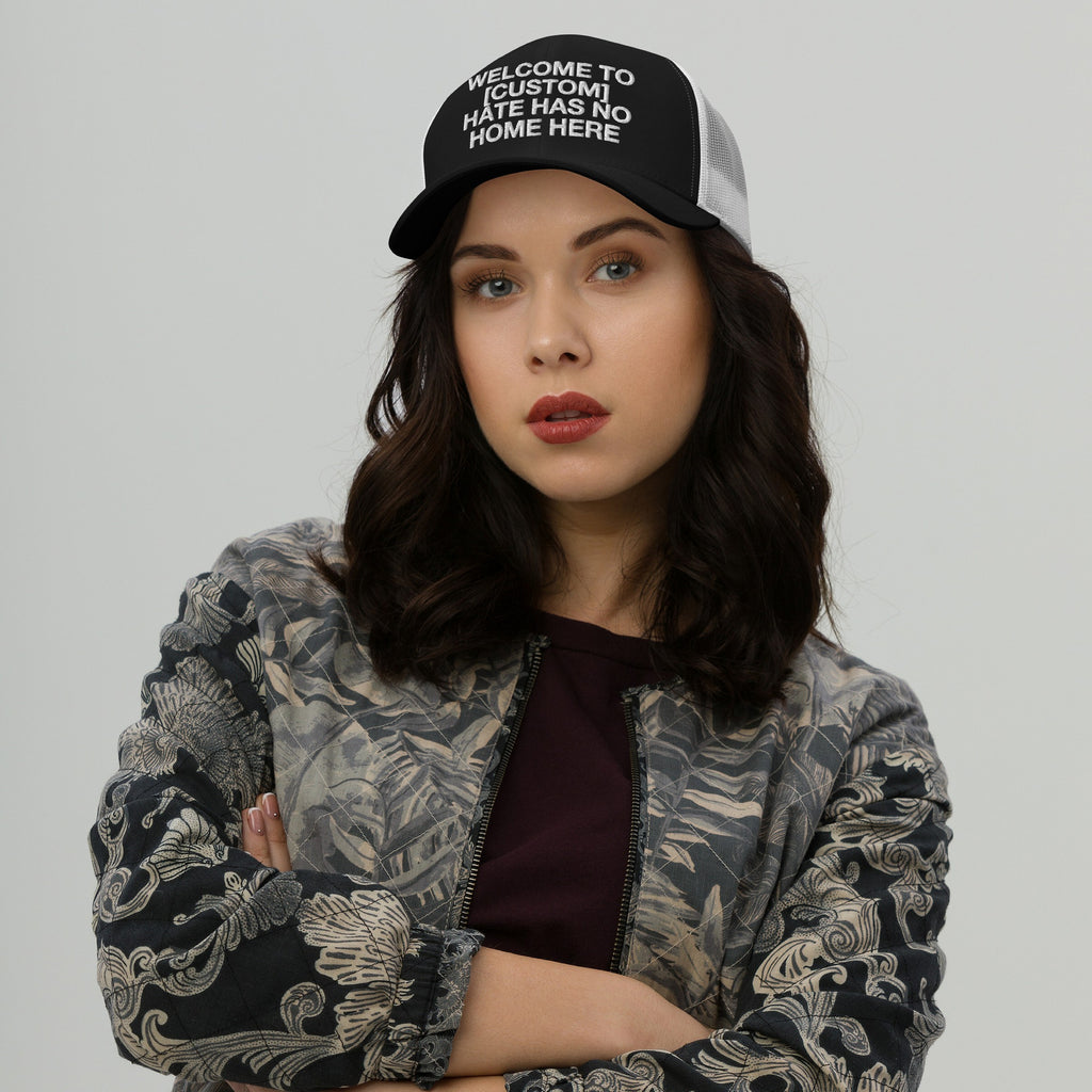 Young woman wearing a Custom Hate Has No Home Here Trucker Hat with a bold slogan, paired with a patterned bomber jacket and burgundy top, exuding confidence and solidarity against hate.