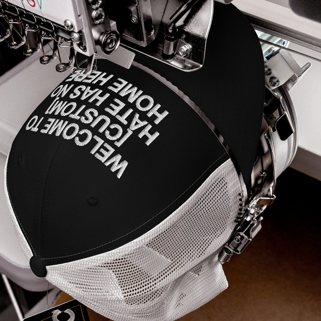 Custom Hate Has No Home Here Trucker Hat on embroidery machine, stitching “WELCOME TO [YOUR CITY]: HATE HAS NO HOME HERE.” Features structured front, breathable mesh back, adjustable snapback for personalization.
