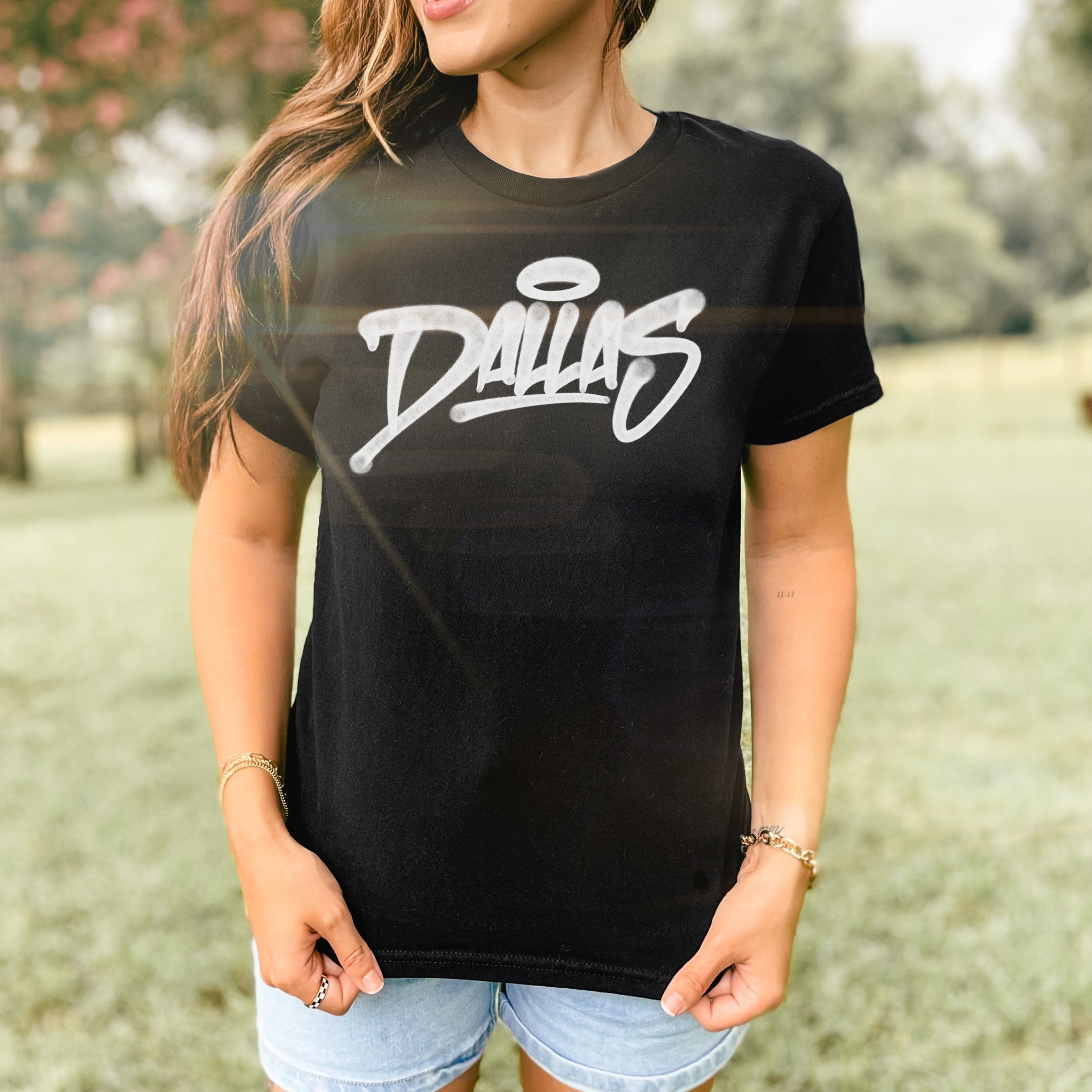 Person wearing Dallas Handstyle Women's Baby Tee outdoors, featuring a black T-shirt with Dallas in white graffiti script and a halo accent, paired with light-wash denim shorts and simple accessories.