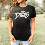 Person wearing Dallas Handstyle Women's Baby Tee outdoors, featuring a black T-shirt with Dallas in white graffiti script and a halo accent, paired with light-wash denim shorts and simple accessories.