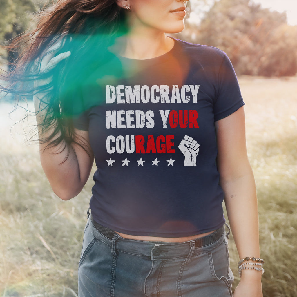 Democracy Women's Baby Tee worn by a young person in a field, showcasing DEMOCRACY NEEDS YOUR COURAGE with highlighted words and a raised-fist graphic, paired with cargo pants and beaded bracelets.