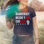 Democracy Women's Baby Tee worn by a young person in a field, showcasing DEMOCRACY NEEDS YOUR COURAGE with highlighted words and a raised-fist graphic, paired with cargo pants and beaded bracelets.