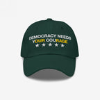 Dark green Democracy Hat with white embroidery: DEMOCRACY NEEDS YOUR COURAGE, highlighting YOUR in yellow, plus five stars below. Ideal for activism and expressing anti-fascist values.