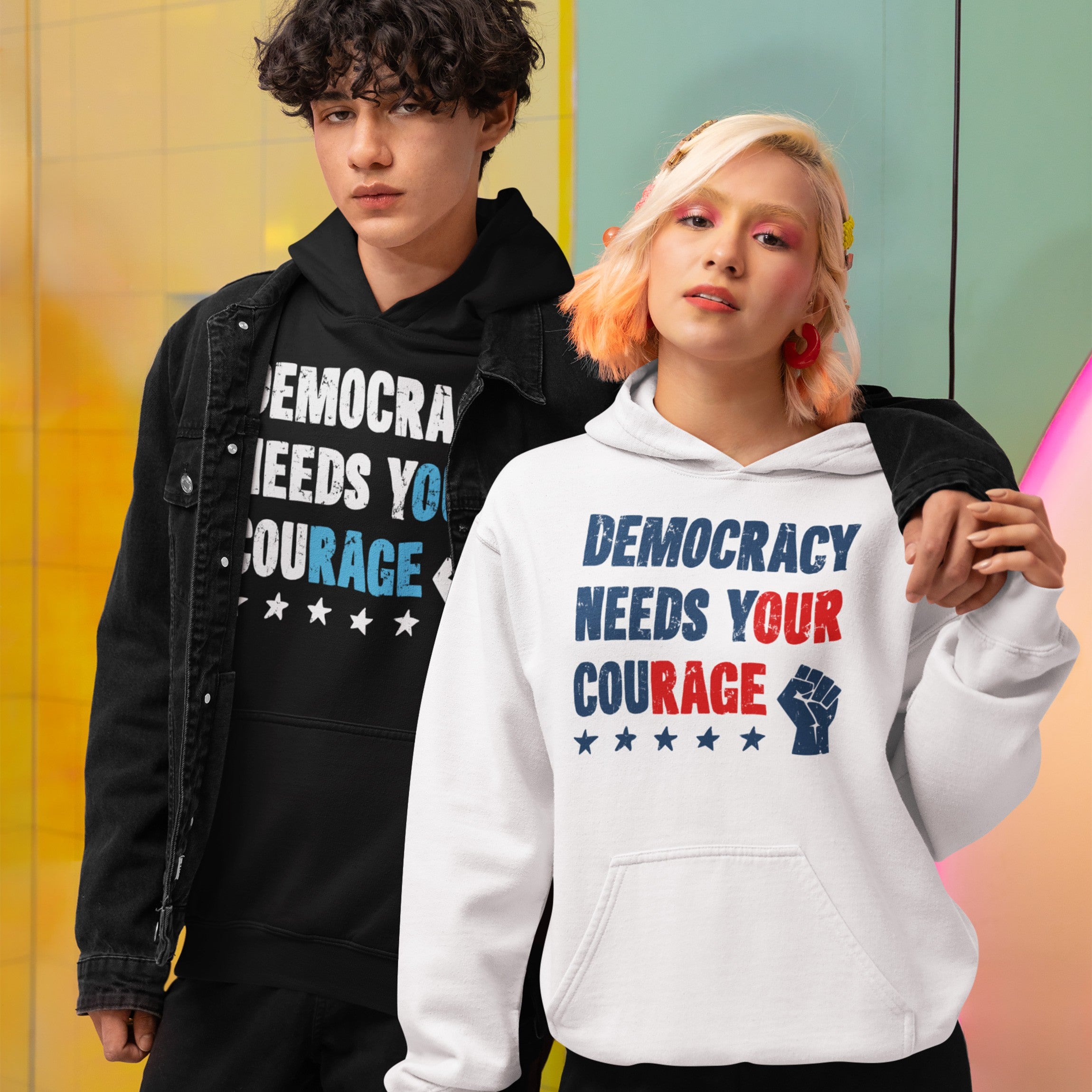 Two young adults showcase the Democracy Shirt, featuring the bold slogan “Democracy Needs Your Courage.” Each hoodie displays protest-inspired graphics that embody resistance and advocacy for equality.