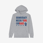 Democracy Shirt: Light-gray hoodie with stencil-style DEMOCRACY NEEDS YOUR COURAGE graphic, featuring distressed stars and raised-fist icon, symbolizing protest energy and resilience. Available in various styles.
