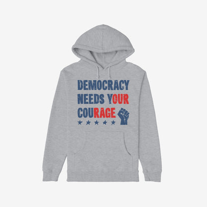 Democracy Shirt: Light-gray hoodie with stencil-style DEMOCRACY NEEDS YOUR COURAGE graphic, featuring distressed stars and raised-fist icon, symbolizing protest energy and resilience. Available in various styles.