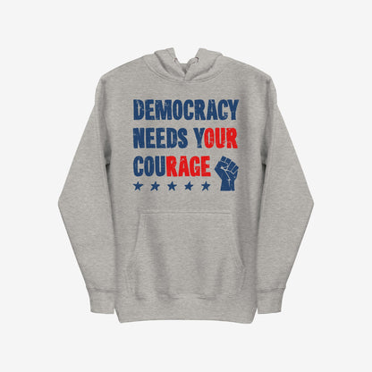 Light-gray Democracy Hoodie features bold “DEMOCRACY NEEDS YOUR COURAGE” text and a blue raised-fist icon, embodying protest spirit. Ideal for standing against injustice in cold weather.