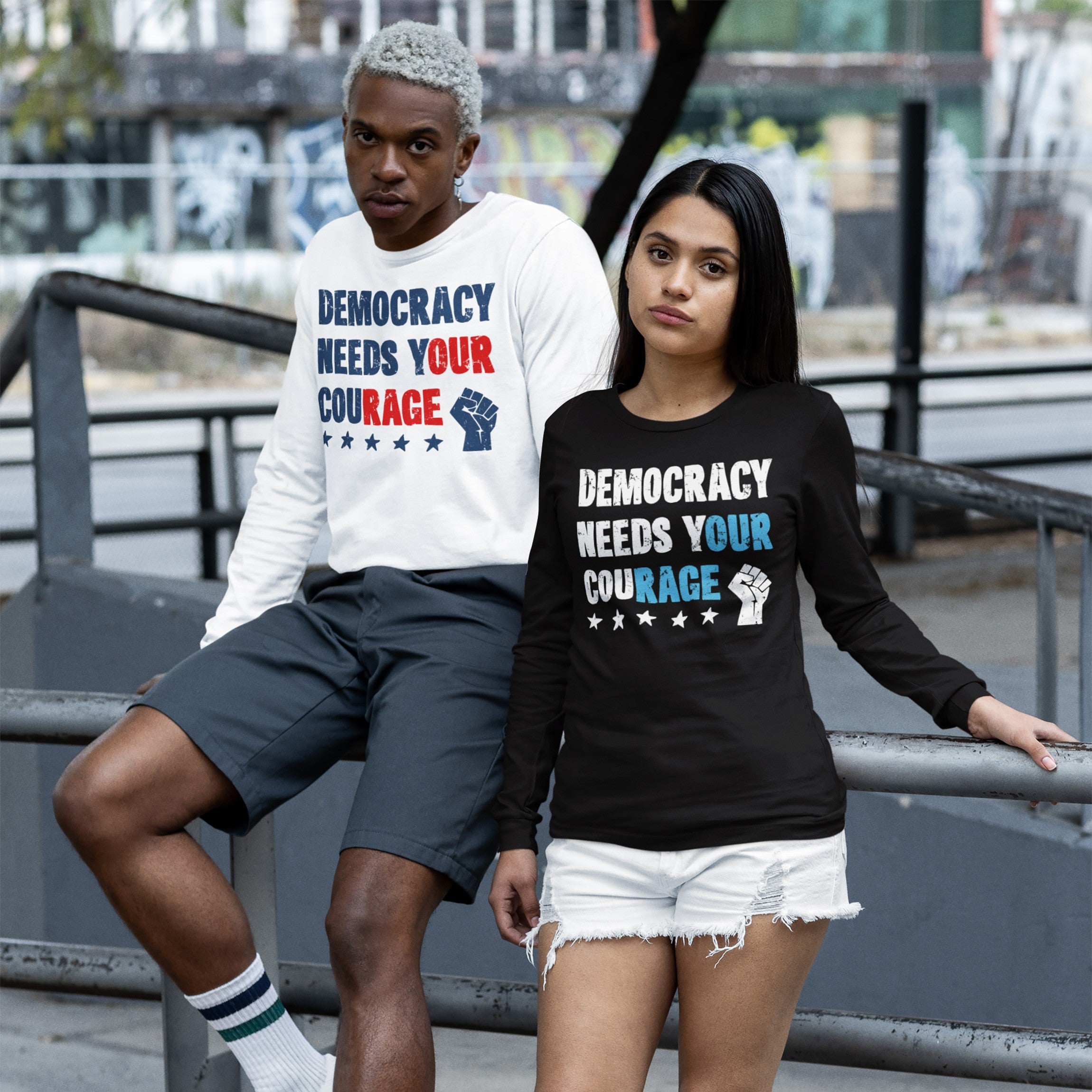 Two young adults wearing Democracy Shirt with Democracy Needs Your Courage slogan, standing in an urban setting. One in white, seated, and the other in black, standing, embodying protest and activism.