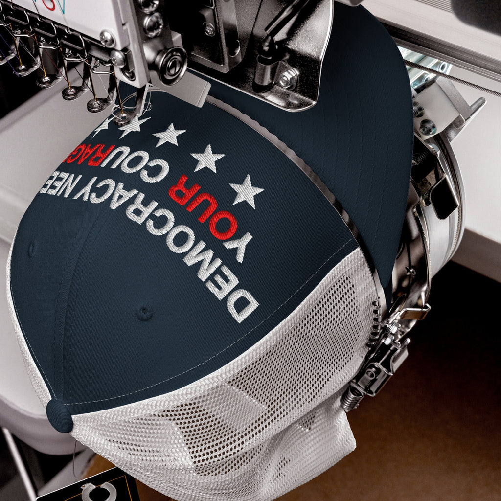 Industrial embroidery machine stitching a Democracy Hat, featuring the slogan “DEMOCRACY NEEDS YOUR COURAGE” in bold, with YOUR highlighted in red, on a navy and white mesh baseball cap.