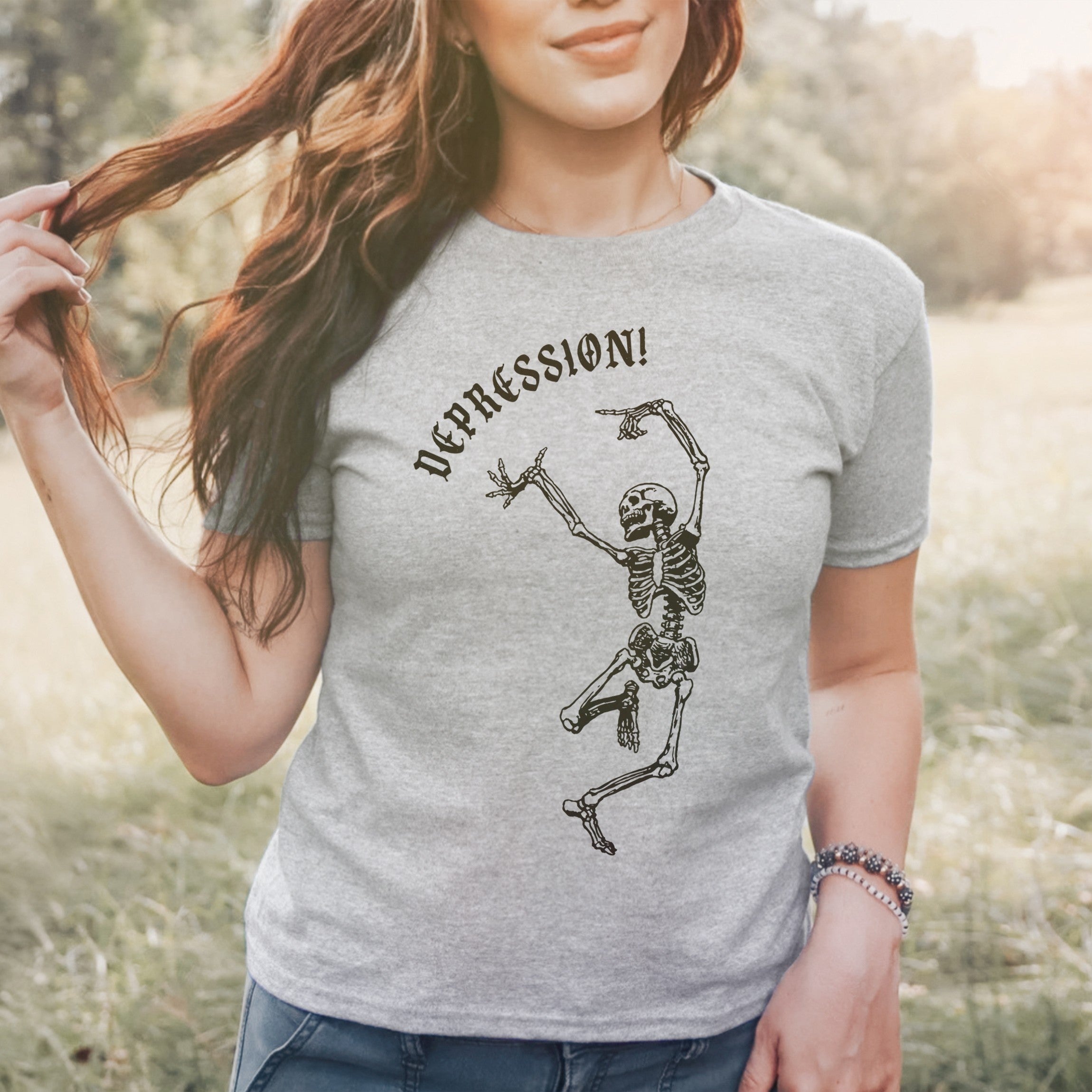 A person outdoors in a light-gray Depression Women's Baby Tee, featuring a dancing skeleton and DEPRESSION! text, embodies stylish self-awareness. Right arm relaxed, left hand lifts wavy hair.