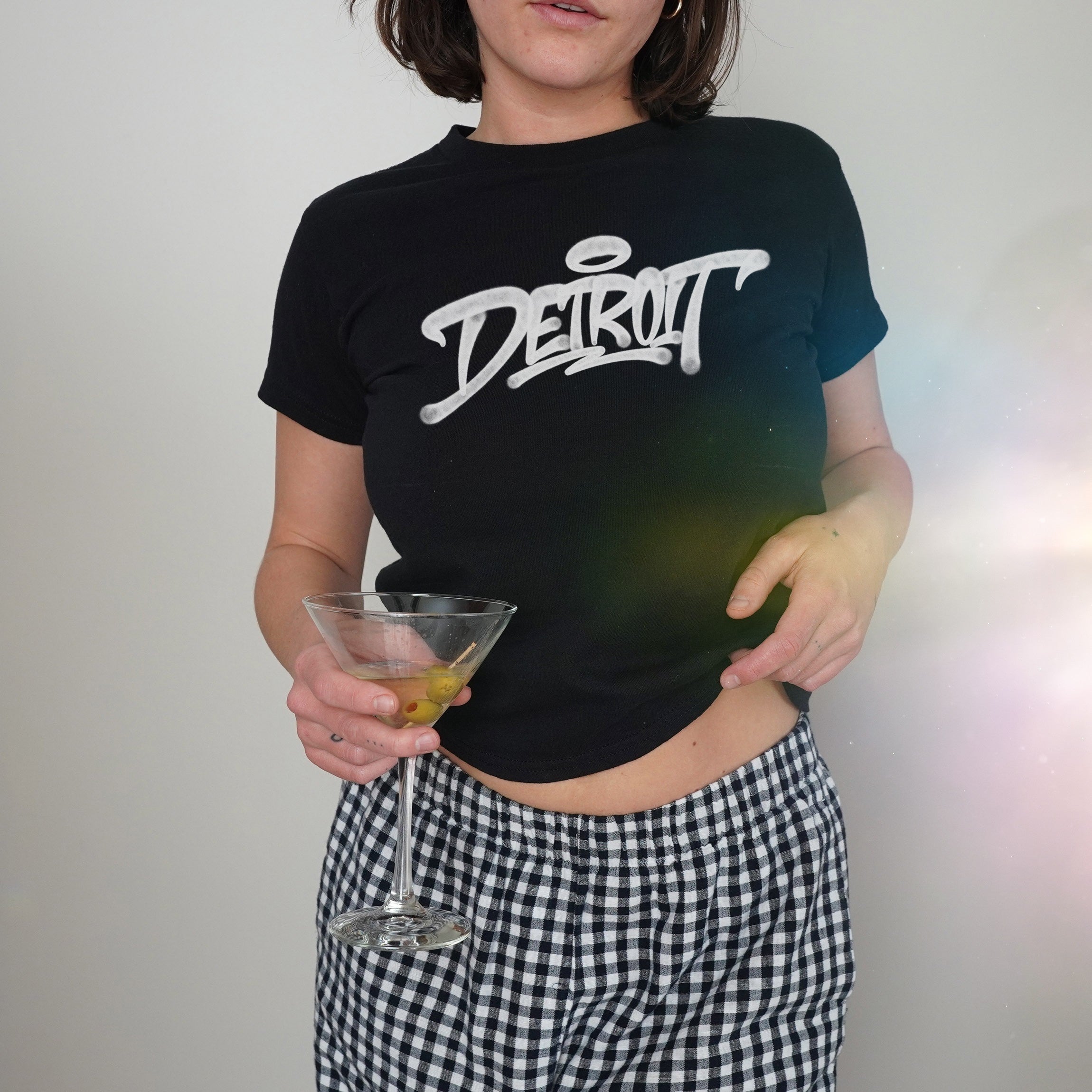 Person wearing Detroit Handstyle Women's Baby Tee, fitted black with Detroit script, paired with checkered pants. They hold a martini glass with olives, slightly lifting shirt hem to reveal midriff.