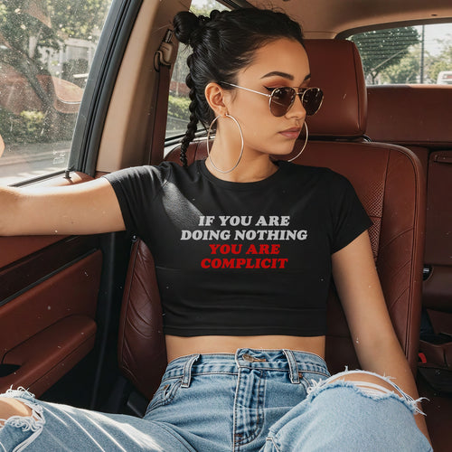 Young woman in a car wears the Don't Be Complicit Baby Tee, featuring a bold slogan. Styled with aviator sunglasses and ripped jeans, this tee embodies protest fashion with a retro Y2K vibe.