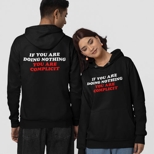 Two models showcase the Don't Be Complicit Hoodie, featuring bold text emphasizing action against complicity. The woman faces forward; the man shows the back, highlighting the statement's impact.