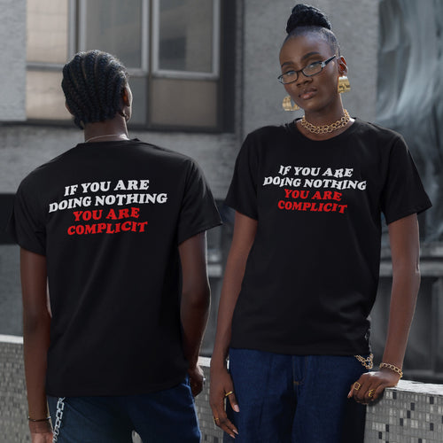 Two young adults model the Don't Be Complicit T-shirt, showcasing its bold white-and-red protest slogan. They wear dark blue jeans and gold jewelry, embodying a modern, statement-driven style.